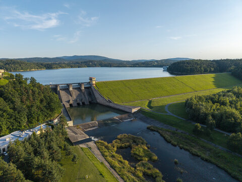 Aerial view of Zapora wodna w Dobczycach dam with a large reservoir, lush green embankments, and surrounding forests under a clear blue sky in Dobczyce, Lesser Poland Voivode, Poland.