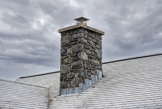 Rugged stone chimney with metal flashing on a grey slate roof against a cloudy sky