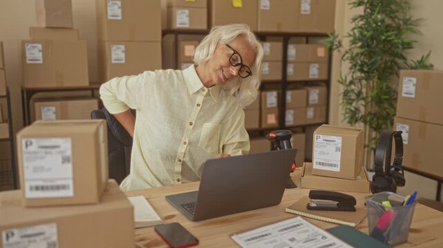 Woman at laptop with hand on lower back amid packed boxes in a shipping building; work fatigue.