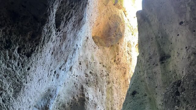 Close upward view inside narrow rocky passage, with porous cliff walls, sunlit beige stone, deep shadow, and rough erosion texture creating dramatic geological contrast in rugged natural setting