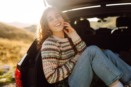 Young woman sits in trunk of a car at sunset in the mountains. Traveler on a road trip, enjoying the mountain scenery, feeling free. Concepts of relaxation, transportation, and enjoyment.