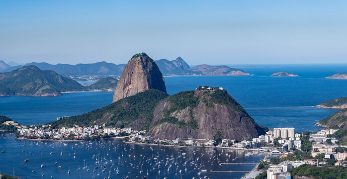 The sweeping view of Sugarloaf Mountain highlights its imposing shape rising above Guanabara Bay, featuring green slopes, blue waters, and the sprawling city. Rio de Janeiro, Brazil.