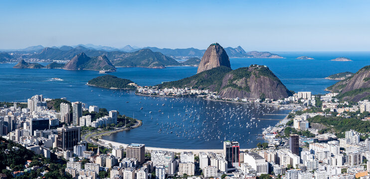 The sweeping view of Sugarloaf Mountain highlights its imposing shape rising above Guanabara Bay, featuring green slopes, blue waters, and the sprawling city. Rio de Janeiro, Brazil.