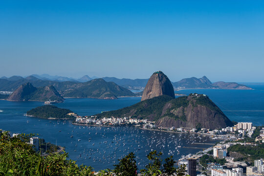 The sweeping view of Sugarloaf Mountain highlights its imposing shape rising above Guanabara Bay, featuring green slopes, blue waters, and the sprawling city. Rio de Janeiro, Brazil.