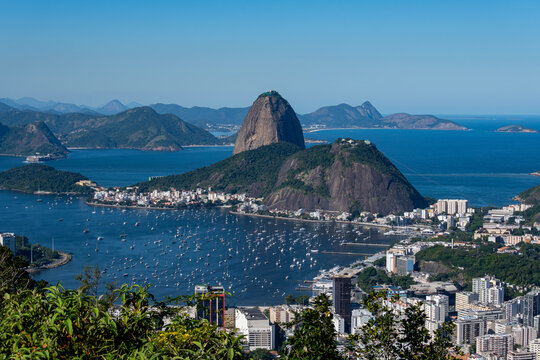 The sweeping view of Sugarloaf Mountain highlights its imposing shape rising above Guanabara Bay, featuring green slopes, blue waters, and the sprawling city. Rio de Janeiro, Brazil.