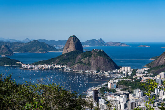 The sweeping view of Sugarloaf Mountain highlights its imposing shape rising above Guanabara Bay, featuring green slopes, blue waters, and the sprawling city. Rio de Janeiro, Brazil.