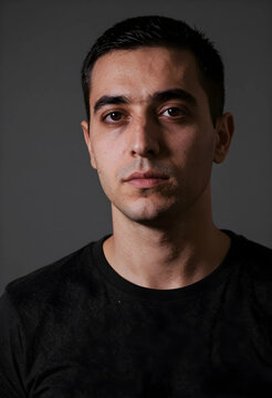 Young adult man with short dark hair and a serious expression looking directly at the camera against a gray background, He wears a black t-shirt against a plain gray studio background