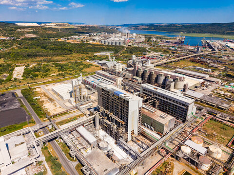View of industrial complex located near a river surrounded by green land and mountains under a clear blue sky during daylight hours