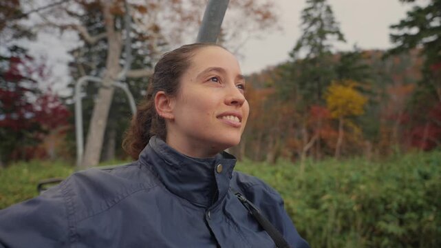 Young smiling woman riding a chairlift up the mountain during autumn, looking at the colorful forest scenery of Daisetsuzan Sounkyo Kurodake Ropeway in Hokkaido, Japan with joy