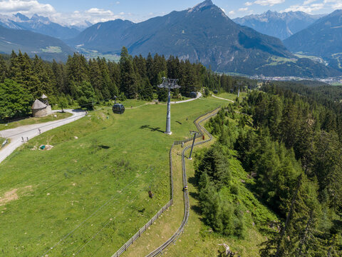 Aerial view of a cable car and a summer toboggan run winding through green alpine meadows and pine forests in the mountains of Imst, Tyrol, Austria.