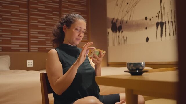Happy woman sitting cross legged on a chair and smiling while browsing social media on her mobile phone in a beautiful traditional japanese style hotel room with tatami mat floor