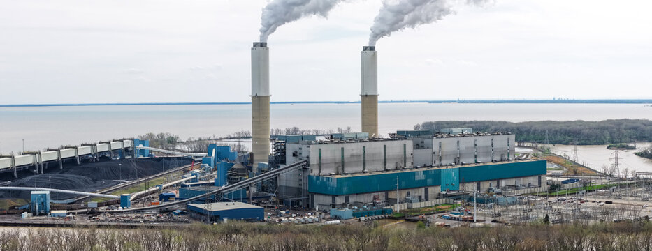 Aerial view of the Monroe Power Plant with two tall chimneys emitting smoke, industrial structures, coal piles, and power lines along the waterfront in Monroe, Michigan, United States.