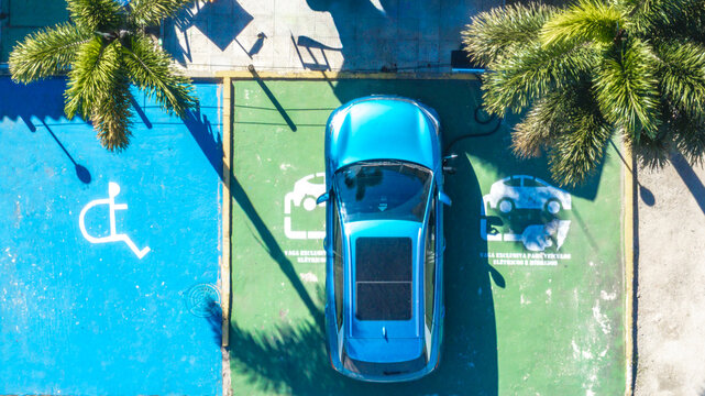 Aerial view of a blue electric car parked in a designated green charging spot with palm tree shadows in Brazil.