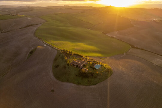 Aerial view of a farmhouse with a swimming pool and cypress trees set in the rolling hills of the Crete Senesi landscape during golden hour in Asciano, Tuscany, Italy.