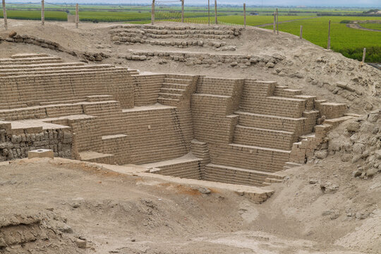 Ancient adobe brick stepped platforms and excavated ruins of a Moche huaca at the El Brujo archaeological complex with green agricultural fields in the background