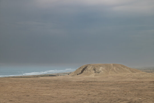 Wide panoramic view of the Huaca Cortada sandy mound in the desert landscape with the Pacific Ocean and overcast sky in the background at El Brujo, Peru