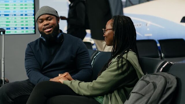 Young Black lovers laugh and chat about an upcoming vacation while cuddling in an airport lounge, excited about travel abroad and bucket list destinations.