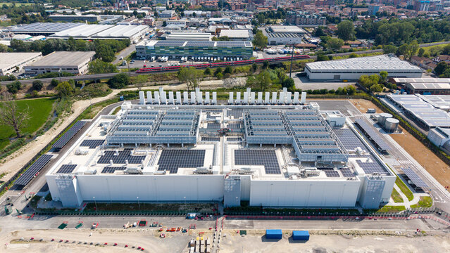 Aerial view of a modern hyperscale data center with extensive rooftop solar panels and cooling infrastructure, located in an industrial area near a railway line in Melegnano, Lombardy, Italy.