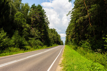 Fototapeta premium Asphalt road stretching ahead through a lush dense green forest on a bright sunny day with scattered clouds in the blue sky creating a scenic perspective for travel