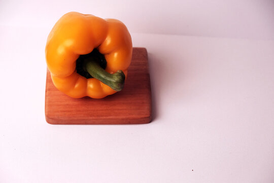 Bright yellow pepper rests on wooden board. Smooth surface gleams under clean white light. Green stem stands tall, contrasting vivid hue. Simple composition draws focus to fresh produce