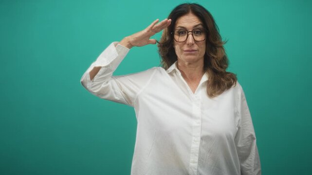 Middle age woman salutes with hand to forehead in teal studio wearing white shirt and glasses; respect dignity.