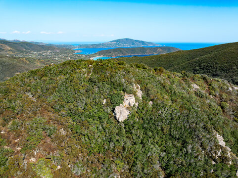 Aerial view of Passo del Monumento surrounded by dense Mediterranean scrubland, rocky outcrops, and the distant coastline of the Tyrrhenian Sea in Campo nell'Elba, Toscana, Italy.