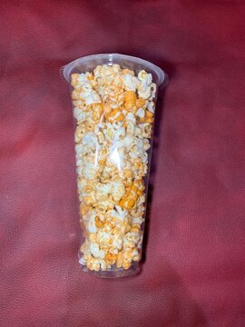 Clear plastic cup filled with seasoned popcorn like cheese or caramel dusted sealed at the top, textured dark red backdrop