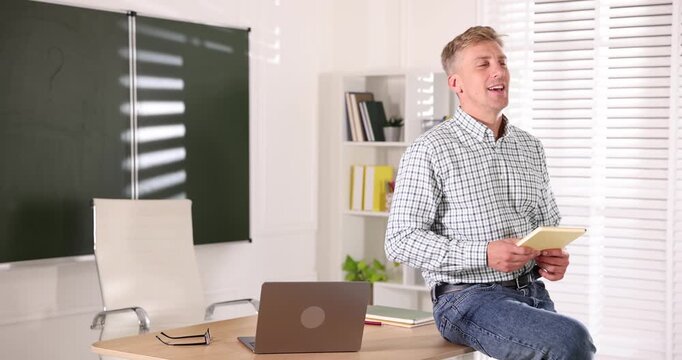 Smiling teacher with book conducting lesson near table in classroom