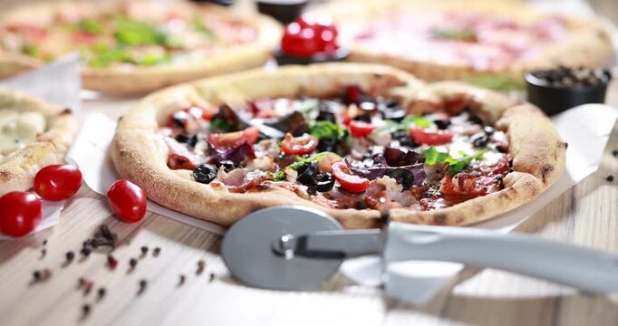 Different yummy pizzas, ingredients and cutter on wooden table, closeup. Camera moving