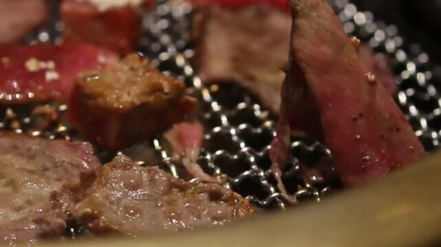 Close-up video footage showing marinated beef and steaks grilling on a hot barbecue, sprinkled with sesame seeds and flipped with tongs at an Asian-style barbecue restaurant.