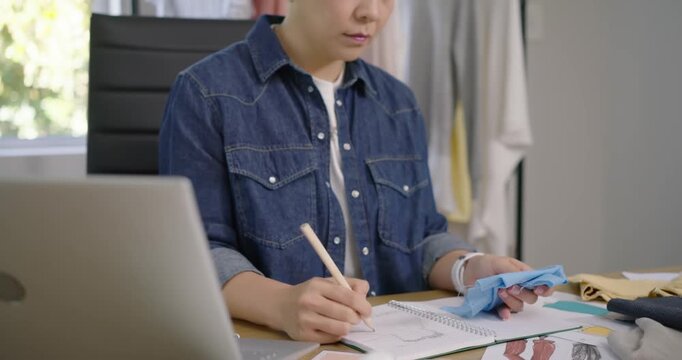 Asian woman sketching in sketchbook holding blue swatch while consulting references for materials