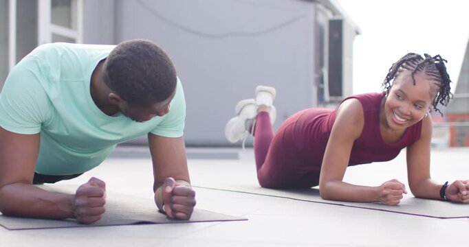 African friends planking on rooftop mats in workout wear, woman entering, smiling and encouraging
