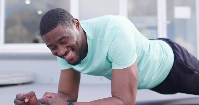 African man spotting offcamera, planking on roof mat, raising head, turning left, smiling, tracker