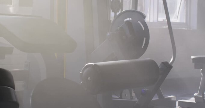 Camera is panning as window light is clearing haze, showing leg curl machine at gym