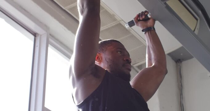 Mid-adult African man gripping pull-up bar and performing pull-ups for strength in bright gym