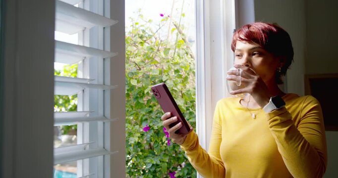 African American woman standing by window holding phone and mug, reading amusing content, smiling