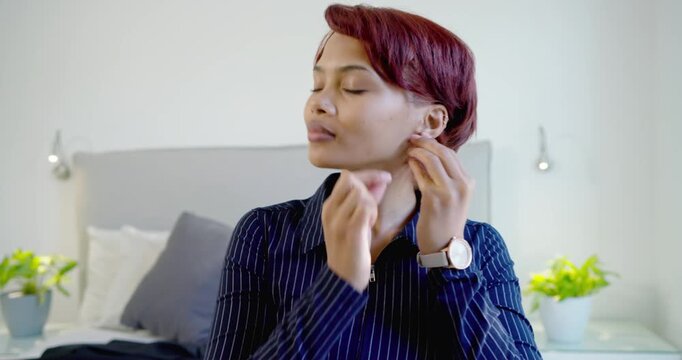 African American woman mid-20s sitting on bed holding earring, inserting earrings, getting ready