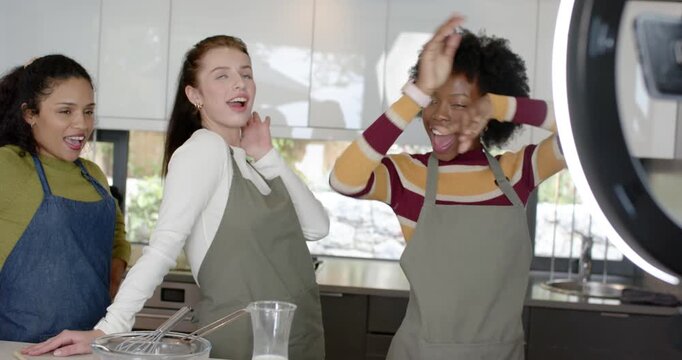 Diverse women in aprons filming cooking demo in kitchen, prompted by smartphone on ring light