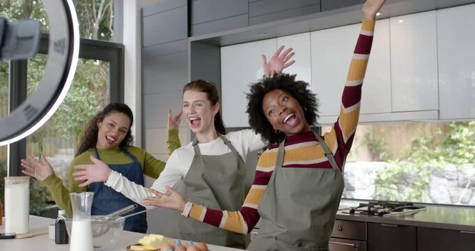 Female friends in aprons doing moves rightmost stepping forward for camera at kitchen island whisk