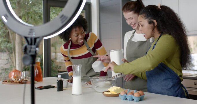 Diverse female friends filming at kitchen island, ring light moving, pouring flour into mixing bowl