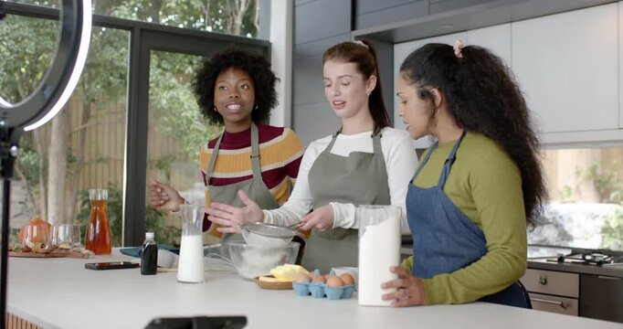 Female friends arranging then sifting flour with sifter for baking at kitchen island with aprons