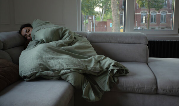 Young woman resting peacefully with closed eyes while wrapped in a warm duvet on the sofa. Comfortable home lifestyle, fatigue, sleep, relaxation and self-care concept.