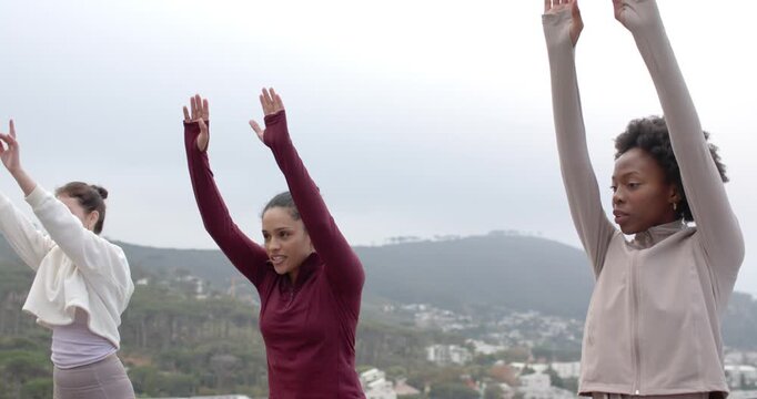 Diverse female friends after cue, practicing forward fold by lowering arms on rooftop yoga mats