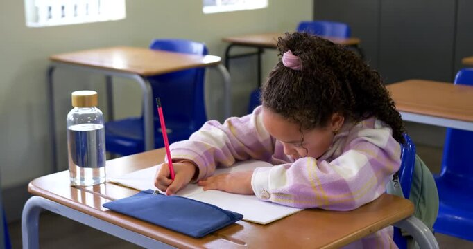 African American schoolgirl writing in notebook with red pencil while doing classwork fidgeting