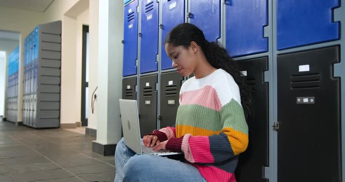 Teen girl in sweater sitting in school hallway by lockers opening laptop and studying with backpack