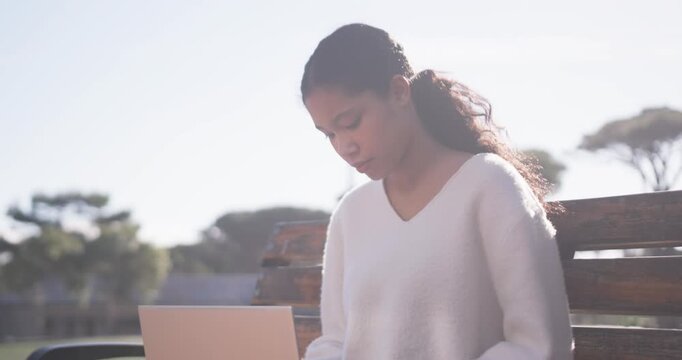 African American child typing on rose-gold laptop on wooden park bench, V-neck, pausing, gazing sky