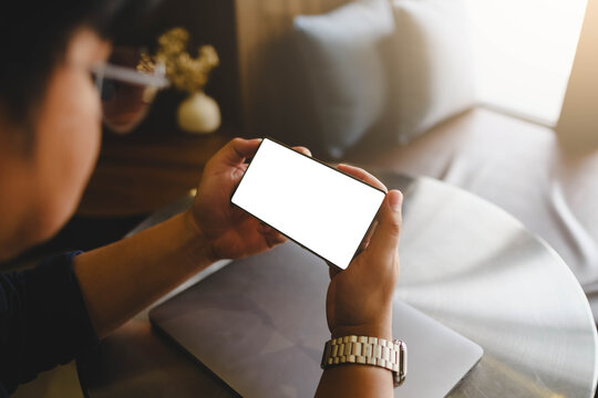 Person holding modern smartphone horizontally with blank white screen mockup over laptop at cafe table.