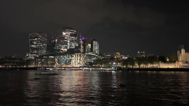 Static nighttime view of the London skyline from the River Thames, featuring The Shard, the Tower of London and shimmering reflections on the water.