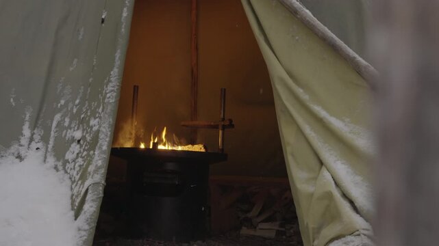 A fire burns steadily in a metal stove inside a campsite tent, surrounded by falling snow. The canvas flaps are partially open, allowing the warm glow of the flames to shine through