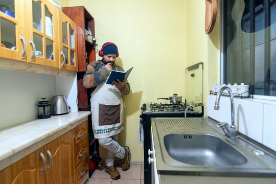 Man in an apron studying a recipe book while listening to music with headphones at home, horizontal shot